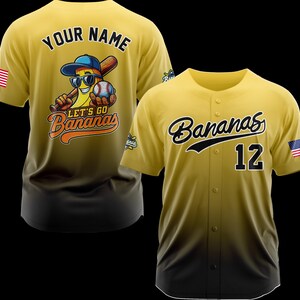 Puede incluir: Dos camisetas de b&eacute;isbol amarillas. Una presenta un personaje de pl&aacute;tano con gafas de sol, un bate de b&eacute;isbol y el texto "Let's Go Bananas". La otra camiseta tiene "Bananas" en escritura y el n&uacute;mero 12. Ambas tienen la bandera americana.