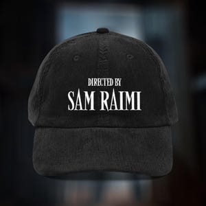May include: Black corduroy baseball cap featuring the text "DIRECTED BY SAM RAIMI" in white. The cap has a curved peak and a button on top. A fashionable accessory for fans of the director.