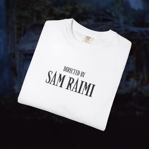 May include: White t-shirt with the text "DIRECTED BY SAM RAIMI" in black, stylized font. The shirt is folded and appears to be made of a soft material. The background is blurred, suggesting an outdoor setting.