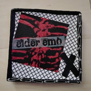 May include: A square decorative plaque featuring a black and white grid background with a central red and black abstract design. The text "elder emo" is printed across the center. The border is adorned with silver safety pins and a black X.