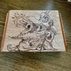 May include: Wooden box with a pyrography design of a wizard holding a staff. The wizard is wearing a hat with a feather and a long robe. The box is rectangular with a hinged lid and a light brown finish.