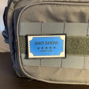 May include: A gray tactical bag with a black zipper and a blue and white patch that reads "SHIT SHOW" with four stars and "ADMIT ONE". The patch is rectangular with a black border and is attached to the bag with velcro.
