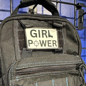 May include: Black tactical bag with a white patch that reads "GIRL POWER" in bold, black letters. The patch also features a gun silhouette and a target symbol. The bag has a zipper closure and light blue stitching.