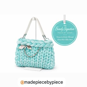 May include: A light blue, chunky-knit handbag with white handles and silver-tone hardware. The bag features a braided top and a decorative charm. A tag reads "The Sandy Signature" and "Grace in Every Thread. Every Piece Tells a Story."