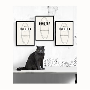 Op de afbeelding: Drie ingelijste prints met het woord "EGGSTRA" in zwarte tekst, boven een grijs ovaal ontwerp, en een zwarte kat die eronder zit. De prints staan op een witte muur. Een zwart-witte paastekening staat op de tafel.