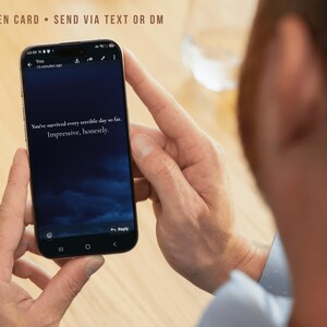 Op de afbeelding: Een smartphone met een donkerblauwe achtergrond en de witte tekst: You've survived every terrible day so far. Impressive, honestly. Bovenaan het scherm staat de tekst: SCREEN CARD • SEND VIA TEXT OR DM. Het apparaat wordt in handen gehouden.
