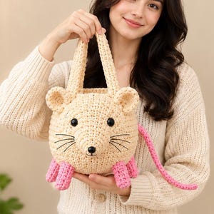 May include: A beige crochet handbag shaped like a mouse with two top handles. The mouse features round ears, black button eyes, a small black nose, and embroidered whiskers. It has four pink crochet legs at the base and a long pink crochet tail.