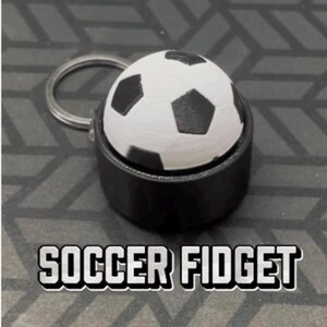 Può includere: Un portachiavi fidget a forma di pallone da calcio bianco e nero inserito in una solida base cilindrica nera con un anello in metallo argentato. Il testo SOCCER FIDGET è stampato in grassetto bianco al di sotto dell'oggetto.