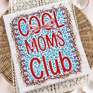 以下が含まれることがあります： 赤い「COOL MOMS CLUB」の文字とリボンが付いた白いTシャツ。テキストは青いヒョウ柄の背景に配置されています。Tシャツは、白いレースのトリムが付いた籐のトレイの上にあります。