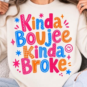 Puede incluir: Una sudadera de cuello redondo color crema con un estampado gráfico multicolor que dice Kinda Boujee Kinda Broke. El diseño incluye varios patrones como lunares, cuadrículas y rayas en tonos rosa, azul y naranja.