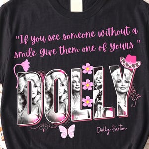 May include: A black t-shirt featuring the text "If you see someone without a smile give them one of yours" in pink. The word "DOLLY" is printed in large letters containing black and white photographs of Dolly Parton, accented with pink flowers, a butterfly, and a pink cowboy hat.