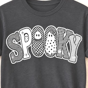 May include: A dark charcoal gray short-sleeve t-shirt featuring the word SPOOKY printed in large white letters. The letters contain various black and white patterns including gingham, polka dots, and stripes, with two ghost shapes integrated.