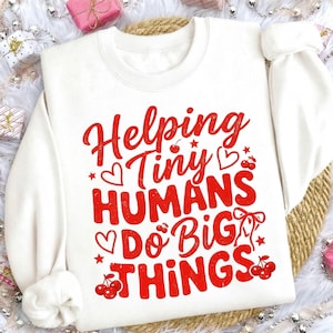 May include: White sweatshirt with the red text "Helping Tiny Humans Do Big Things." The design includes hearts, stars, and cherries. The sweatshirt has long sleeves and a crew neck.