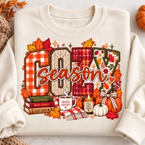 May include: A cream-colored sweatshirt featuring a graphic with the text COZY season. The design includes large letters filled with plaid and knit patterns, surrounded by autumn leaves, a stack of books, a mug labeled SWEATER WEATHER, a candle labeled FALL FEELS GOOD, and pumpkins.