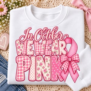 Op de afbeelding: Een wit sweatshirt met ronde hals en de tekst In October We Wear Pink in een groot lettertype met diverse patronen. De letters zijn gevuld met roze gingham-, bloemen- en stippenpatronen, vergezeld door een roze lint met een strik.