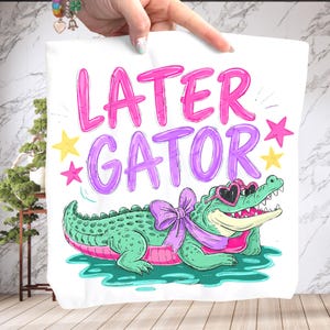 Peut inclure: Tissu blanc avec l'inscription « LATER GATOR » en rose et violet. Un alligator de dessin animé portant des lunettes de soleil en forme de cœur et un nœud est en dessous du texte. L'alligator est vert avec un ventre rose et est dans l'eau.
