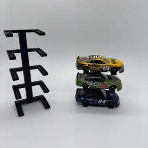 May include: A black plastic vertical display rack with five tiers and three die-cast stock car models stacked on the right. The top car is yellow with number 20, the middle is green with number 54, and the bottom is blue with number 84.