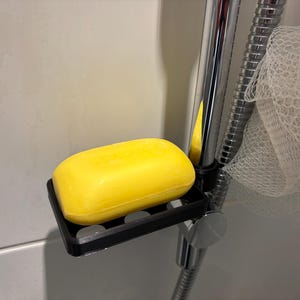May include: A bright yellow bar of soap rests on a black plastic soap dish attached to a chrome shower head. The soap dish has a rectangular shape with drainage holes. A white mesh sponge hangs nearby.