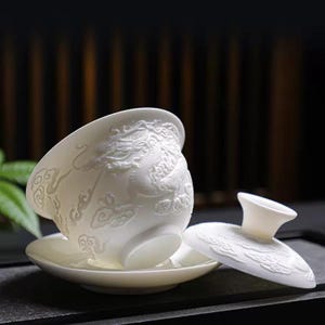 Op de afbeelding: Een witte porseleinen theeset met een kopje, schotel en deksel. De kop en het deksel zijn versierd met een gedetailleerd drakenreliëf. De set staat op een donkere ondergrond, met een vage achtergrond.