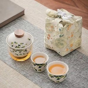 May include: A tea set comprising a lidded ceramic cup with a wooden lid, two small teacups, and a floral-patterned bag. The cups and lidded cup are adorned with white flowers and green leaves. The tea inside is amber-coloured.