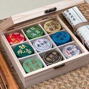 May include: A wooden box with a hinged lid, containing nine small, round tea cakes of various colours. Each cake has Chinese characters printed on it. A scroll with Chinese text is visible in the background.