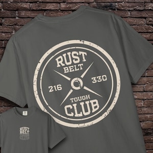 Op de afbeelding: Donkergrijs t-shirt met een verweerde witte afbeelding. De afbeelding toont de woorden "RUST BELT TOUGH CLUB" rond een gewichtplaatontwerp, met de nummers 216 en 330. Een kleinere versie van het ontwerp is onderaan zichtbaar.