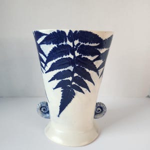 May include: A white ceramic vase with a flared top and a wide base. The vase is decorated with a large blue fern leaf design. Small, decorative blue handles are on either side of the vase.