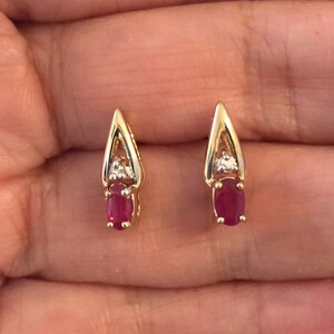 May include: A pair of gold drop earrings featuring a pointed triangular frame. Each earring is set with a small, clear, faceted stone at the top and a vibrant, oval-shaped, deep red gemstone at the base, secured by four metal prongs.