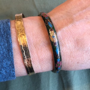 Op de afbeelding: Twee armbanden op een pols. Eén is een gehamerde goudkleurige metalen armband. De andere is een donkergroene emaille armband met een bloemmotief in roze, blauw en goud. De armbanden worden om een pols gedragen.