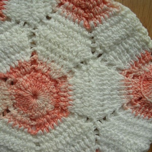 Puede incluir: Primer plano de un artículo de ganchillo hecho a mano con una combinación de colores blanco y coral. La pieza tiene un patrón geométrico con un diseño floral central. La textura es visible, mostrando los bucles y nudos del hilo. Se adjuntan dos lazos de hilo blanco.