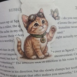 May include: A magnetic bookmark featuring an illustration of a brown tabby kitten wearing aviator goggles and a green collar with a circular charm depicting a tree. The kitten is raising one paw toward a small butterfly flying to its right.