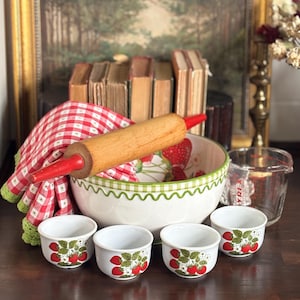 May include: A large white ceramic mixing bowl with a green checkered rim and strawberry pattern, four matching small bowls, a wooden rolling pin with red handles, a red and white checkered cloth, and a clear glass measuring cup.