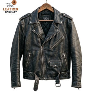 May include: A black distressed leather motorcycle jacket featuring a quilted interior lining, silver-tone metal zips, a snap-button flap pocket, and an adjustable waist belt. The inner label reads The Vintage Leather Co. The jacket has epaulettes.
