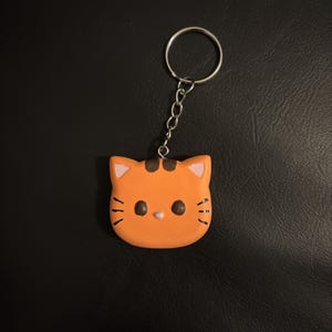 May include: Orange cat-shaped keychain with a silver keyring and chain. The cat face has black whiskers, brown eyes, and a pink nose and ears. The keychain is a cute accessory.
