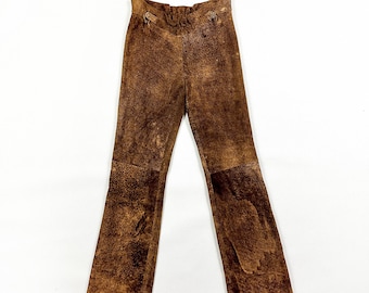 May include: Brown suede pants with a flared leg design. The pants have a high waist and two decorative buttons. The suede has a distressed look with variations in color.