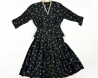May include: A vintage black two-piece suit with a skirt and a button-down top. The suit features a pattern of small, scattered gold, white, and brown dashes. The top has a cream-colored collar and three-quarter sleeves. The skirt is pleated.