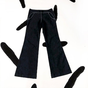 May include: Dark wash denim trousers with a flared leg design. The trousers feature white stitching details along the pockets and seams. The trousers are displayed on a white background with black abstract paint strokes.