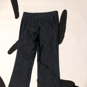 May include: Dark blue denim pants with a straight leg design. The pants have a high waist and a zipper closure. White stitching accents the seams. The pants are displayed on a white background with black abstract shapes.