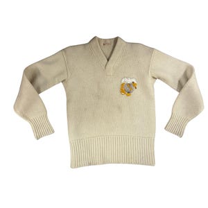 May include: Cream-colored vintage V-neck sweater with ribbed cuffs and hem. Features a small embroidered patch with a globe and bear design in yellow, brown, and white. The sweater has long sleeves and a relaxed fit, perfect for layering.