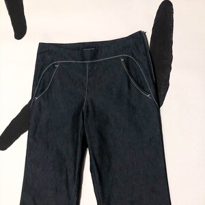May include: Dark blue denim pants with white stitching detail. The pants have a straight leg cut, a zipper closure, and curved front pockets. The brand name is visible on the waistband. The pants are displayed against a white background with black abstract shapes.