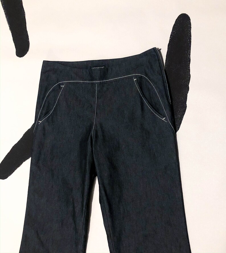 May include: Dark blue denim pants with white stitching detail. The pants have a straight leg cut, a zipper closure, and curved front pockets. The brand name is visible on the waistband. The pants are displayed against a white background with black abstract shapes.