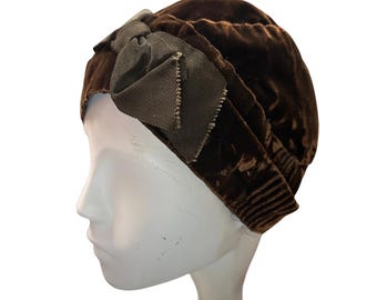 May include: A brown velvet turban-style hat with a decorative bow. The hat is a rich, dark brown color and has a soft, textured appearance. The bow is a contrasting shade of brown. The hat is displayed on a white mannequin head.