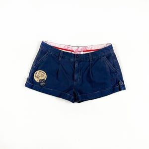 May include: Dark blue denim shorts with a pink and red waistband. The shorts have a patch on the left leg with the text "University of Pink" and a crest with the letter "P".