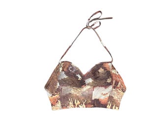May include: A cropped halter top featuring a sweetheart neckline and thin neck ties. The garment is printed with a detailed, multi-colored artistic scene in shades of brown, tan, and cream, depicting architectural structures and human figures.