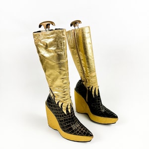 May include: A pair of gold knee-high boots with black crocodile-textured accents. The boots have a wedge heel and a pointed toe. The gold material has a crinkled texture. The boots are displayed on a white background.