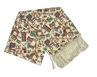 May include: A beige scarf with a repeating pattern of various furniture pieces in brown, green, and blue. The scarf has a cream-colored fringe along one edge. The furniture includes tables, chairs, and lamps.
