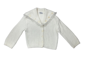 May include: A white knit cardigan with a sailor collar and fringe trim. The button-down sweater has long sleeves and a textured knit pattern. The collar is adorned with small, round buttons.