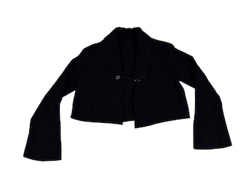 May include: A black cropped jacket with wide, bell sleeves. The jacket has a shawl collar and fastens with a single clasp at the front. The sleeves are long and flared, creating a dramatic silhouette.