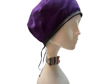 May include: A purple beret with a black leather trim is displayed on a mannequin head. The beret has a classic shape and is made of a textured fabric. Black ties hang from the back of the beret. A silver choker is around the mannequin's neck.