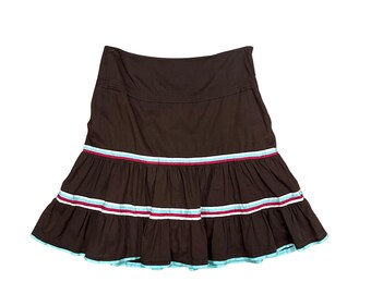 May include: A dark brown skirt featuring a wide waistband and two tiered ruffles at the bottom. The skirt is accented with horizontal stripes in light blue, white, and magenta along the seams of the ruffles and at the hemline.
